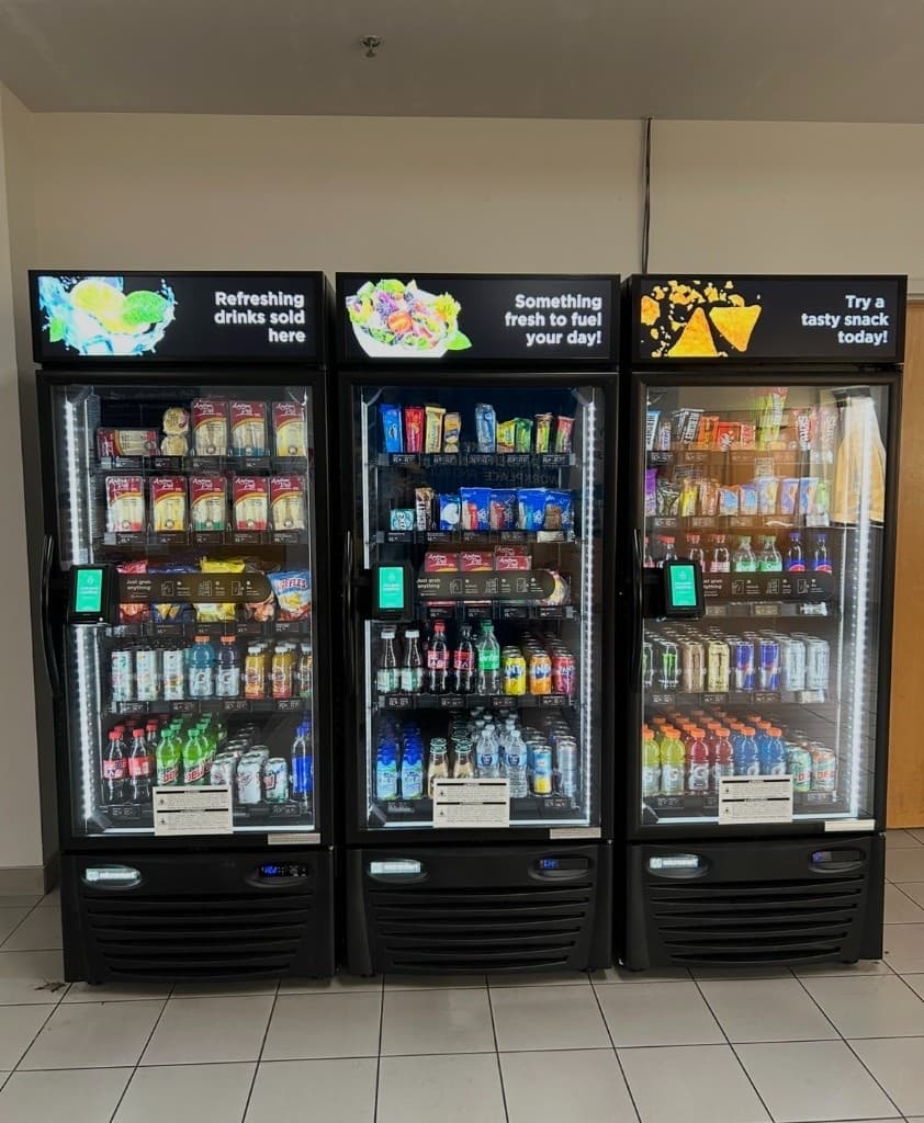 Three stocked AI vending machines with glass doors and payment screens in a Jacksonville warehouse breakroom