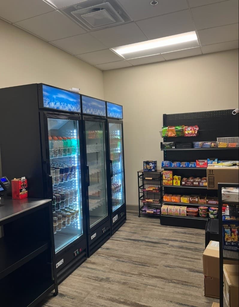Apartment micro-market with three refrigerated glass doors, open snack shelving, and a checkout counter in a compact indoor room